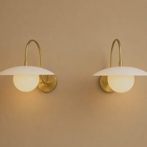 May include: Two wall sconces featuring a curved brass arm, a circular brass wall mount, a wide white disc-shaped shade, and a central spherical white glass globe. The fixtures are mounted side by side, emitting a soft, warm glow from the globes.