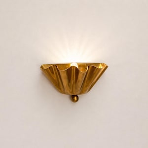 May include: A gold-colored wall sconce with a scalloped, shell-like design. The sconce has a small, round detail at the bottom and emits a warm, inviting light. The fixture is mounted on a white wall.