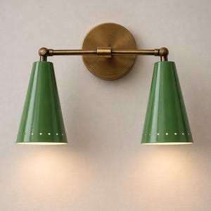 Puede incluir: Aplique de pared con placa trasera circular de latón y brazo horizontal que sostiene dos pantallas verdes en forma de cono. Cada pantalla tiene una fila de pequeños agujeros espaciados cerca de la base. Para uso en interiores.
