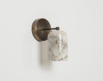 May include: A wall-mounted light fixture with a circular brass base and a cylindrical shade. The shade is made of white marble with brown veining. The fixture is designed for indoor use.