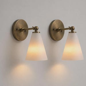 May include: A pair of wall sconces featuring circular antique brass backplates, adjustable horizontal arms, and conical frosted glass shades. Each fixture emits a soft, warm glow, highlighting the metallic finish and the translucent texture of the glass.