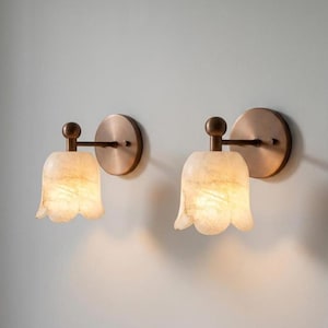 Può includere: Una coppia di applique da parete con piastre circolari color bronzo e bracci orizzontali che sostengono paralumi in pietra bianca traslucida a forma di campana. Ogni lampada presenta un piccolo terminale sferico sulla parte superiore.