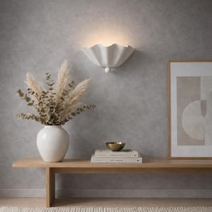 May include: A white, shell-shaped wall light casting a warm glow on a grey wall. Beneath, a wooden bench displays a white vase with dried flowers, books, and a small gold bowl. A framed print adds to the decor.