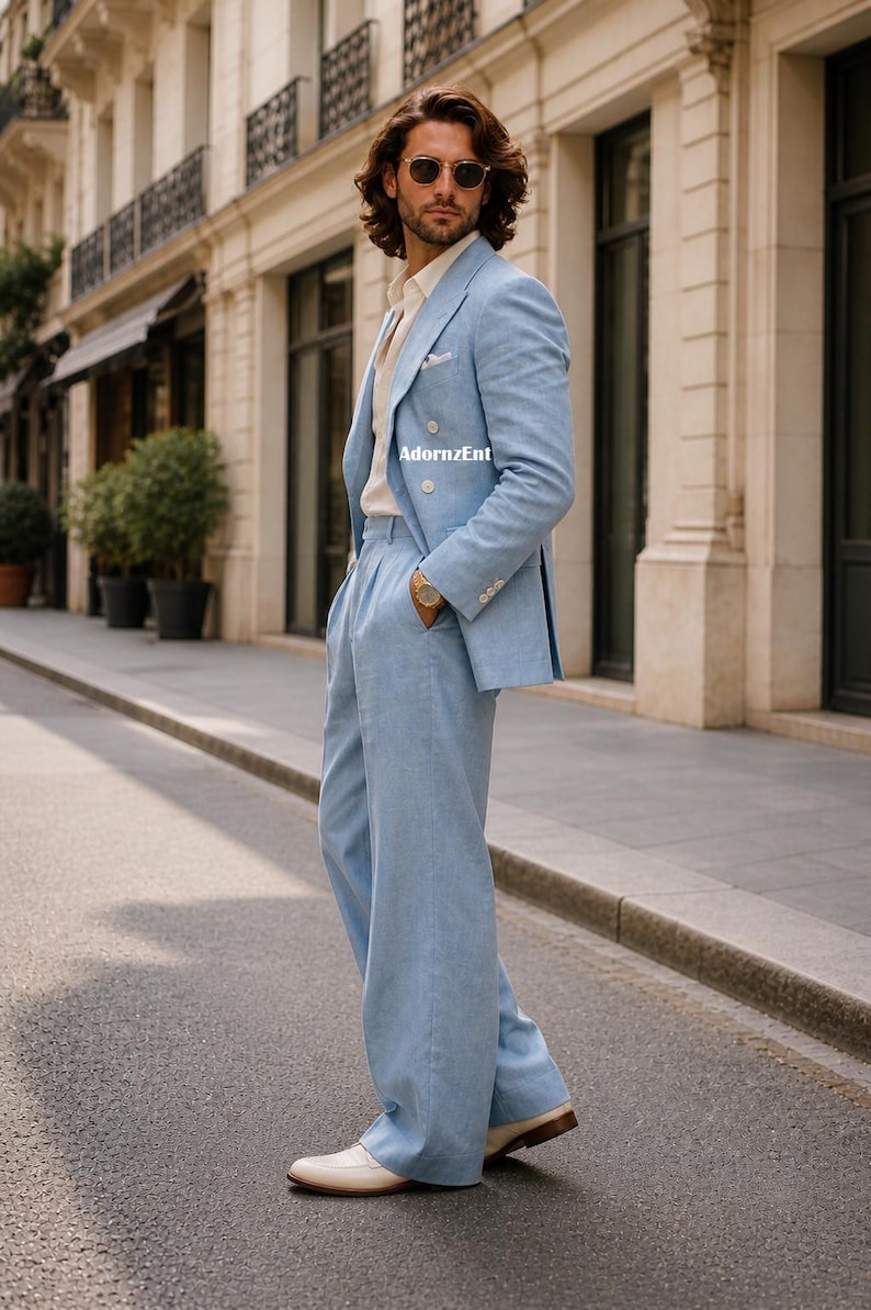 May include: A light blue linen suit with a double-breasted blazer and wide-leg pants. The suit is paired with a cream-colored shirt and white shoes. The image is taken outdoors on a city street.