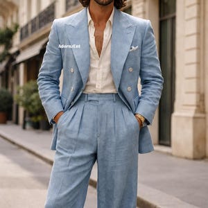 May include: A light blue double-breasted suit with matching wide-leg pants. The suit is worn over a cream-colored button-down shirt. The image is taken outdoors on a city street. The suit has a pocket square and gold watch.