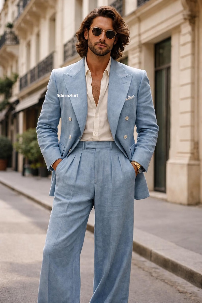 May include: A light blue double-breasted suit with matching wide-leg pants. The suit is worn over a cream-colored button-down shirt. The image is taken outdoors on a city street. The suit has a pocket square and gold watch.