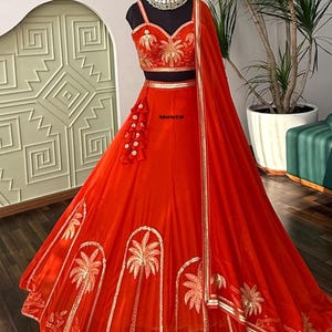 May include: A vibrant orange lehenga set featuring a sleeveless sweetheart neckline blouse and a voluminous floor-length skirt. Both pieces are adorned with gold embroidered palm tree motifs and metallic trim. A matching sheer dupatta is included.