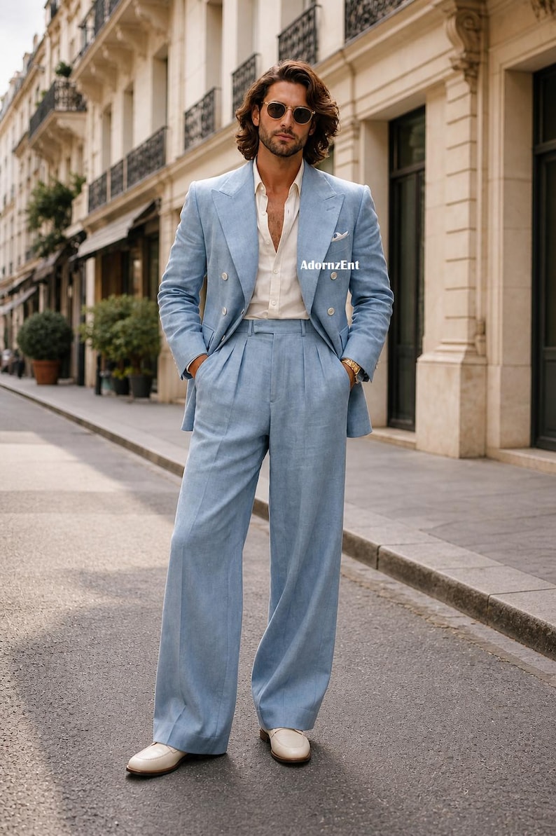 May include: A light blue linen suit, featuring a double-breasted blazer and wide-leg trousers. The suit is paired with a white button-down shirt and white loafers. The outfit is completed with sunglasses and a gold watch. The image is taken outdoors.