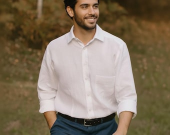 May include: A white button-down shirt with a chest pocket and rolled-up sleeves. The shirt is paired with blue pants and a black belt. The person is walking outside in a natural setting.