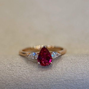 Puede incluir: Un anillo de oro rosa con una gran gema roja oscura en forma de pera como pieza central. Dos piedras más pequeñas y claras en forma de pera flanquean la piedra principal. El anillo está sobre un fondo neutro.
