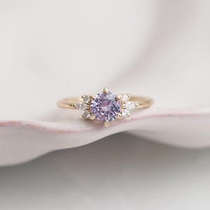 Puede incluir: Un anillo de oro con una piedra central redonda de color púrpura claro, rodeada de piedras blancas más pequeñas. El anillo tiene una banda delgada de oro, con pequeñas piedras blancas a cada lado de la piedra principal. El anillo está sobre una superficie rosa claro.