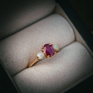 May include: A gold ring with a central oval ruby and two opal side stones. The ring is set in a gold band and is presented in a black jewellery box with a light grey interior.