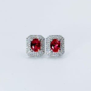 Puede incluir: Un par de pendientes de botón cuadrados. Cada pendiente presenta una gema roja ovalada vibrante, rodeada por un halo de pequeñas piedras blancas brillantes. Los pendientes están colocados sobre un fondo blanco liso.