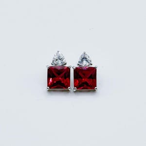May include: A pair of silver-toned stud earrings. Each earring features a square, deep red gemstone below a smaller, triangular clear stone. The earrings are set against a white background.