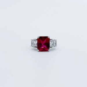 May include: A silver ring with a large, rectangular, ruby-red gemstone at its centre. Smaller, clear, rectangular stones are set either side of the main stone. The ring is displayed against a plain white background.