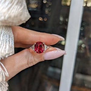 Op de afbeelding: Een zilveren ring met een grote, ovaal geslepen, helderrode edelsteen als middelpunt. Twee kleinere, driehoekige, heldere edelstenen flankeren de hoofdsteen. De ring wordt tegen een vage achtergrond gehouden, wat het ontwerp benadrukt.