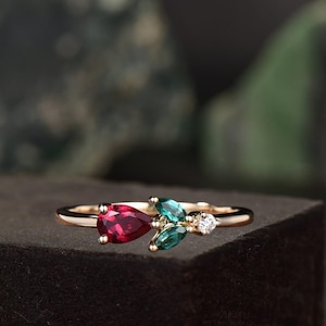 Op de afbeelding: Een gouden ring met een peervormige, dieprode edelsteen, geflankeerd door twee teal-kleurige, bladvormige stenen en een kleine, ronde, heldere steen. De ring heeft een delicate, dunne band.