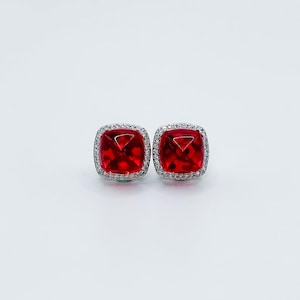 May include: A pair of square-cut, red gemstone stud earrings. Each earring features a vibrant red stone surrounded by a halo of small, clear gemstones set in a silver-colored metal. The earrings are displayed against a white background.