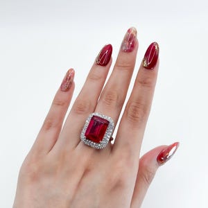 May include: A rectangular ruby ring with a halo of small, clear gemstones. The ring is set on a hand with red nail polish. The ruby is a deep red colour.