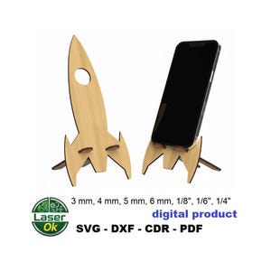 May include: Two wooden rocket-shaped phone stands. One stand is empty, and the other holds a black smartphone. The stands are made of light-colored wood with a natural finish. The image includes text that says "digital product" and "SVG - DXF - CDR - PDF".