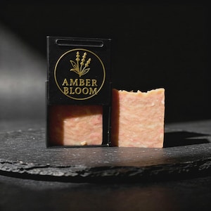 Könnte beinhalten: Ein Seifenstück und seine Verpackung. Die Seife hat eine hellpfirsichfarbene Farbe mit marmoriertem Aussehen. Die Verpackung ist schwarz mit einem goldenen Emblem und den Worten "AMBER BLOOM".