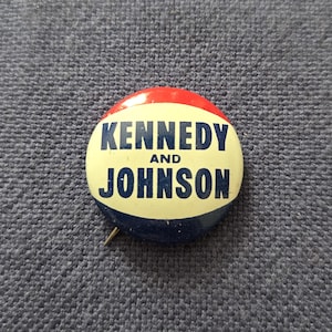 May include: A vintage campaign button with a red, white, and blue color scheme. The button features the text "KENNEDY AND JOHNSON" in navy blue lettering. The pin is round and has a metal pin on the back.