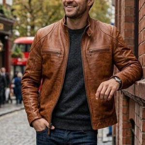 Op de afbeelding: Bruine leren jas met ritssluiting en twee borstzakken. De jas wordt gedragen met een donkergrijze trui en een blauwe jeans. Op de achtergrond een bakstenen gebouw en een geplaveide straat.