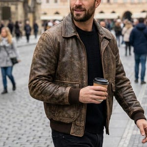 Op de afbeelding: Bruine leren bomberjack met een klassieke kraag en geribde manchetten. De jas wordt gedragen over een zwarte trui en gecombineerd met een zwarte jeans. De persoon houdt een koffiekopje vast en loopt op een geplaveide straat.