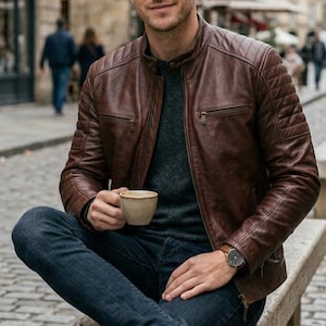 Op de afbeelding: Een bruine leren jas met een gewatteerd ontwerp op de schouders en mouwen. De jas heeft een ritssluiting en een opstaande kraag. De persoon draagt een donkergrijze trui, een blauwe jeans en bruine leren laarzen. Een horloge zit om de pols.