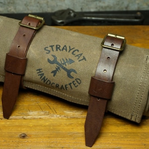 Puede incluir: Un rollo de herramientas de lona enrollado con correas de cuero marrón y hebillas de latón. El rollo es de color canela con las palabras "STRAYCAT HANDCRAFTED" impresas en negro. El rollo de herramientas está sobre una superficie de madera.