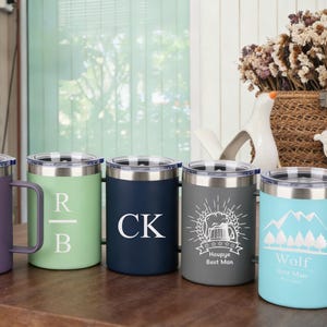 Könnte beinhalten: Fünf Reisebecher aus Edelstahl in verschiedenen Farben, jeweils mit einem anderen Design. Die Becher sind lila, grün, marineblau, grau und hellblau. Zu den Designs gehören eine Berg- und Zahnradgrafik, Initialen und eine Bierkrug-Illustration.