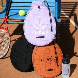 Op de afbeelding: Een rood-zwarte tennisracket, drie tennisracketvormige tassen in zwart, oranje en lavendel, en een waterfles. Op de lavendelkleurige tas staat de naam "Nathan". Een tennisbal ligt op een blauwe bank.