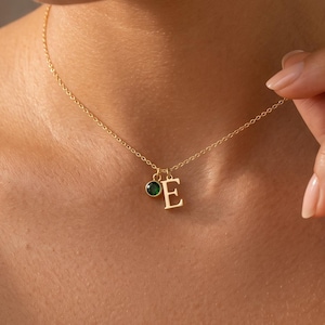 May include: A gold-toned chain necklace featuring a small, round, dark green gemstone charm and a gold-toned letter E pendant. The delicate metal chain rests against the neck, showcasing the two small charms hanging together at the center point.
