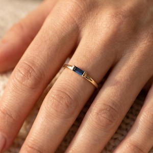 Peut inclure: Une bague en or ornée d'une pierre précieuse rectangulaire bleu foncé. La bague présente un design minimaliste avec un fin anneau. La bague est présentée sur une main, avec un pull de couleur neutre en arrière-plan.