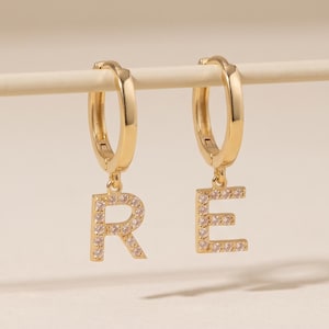 May include: A pair of gold-toned hoop earrings, each featuring a dangling letter charm. The left earring displays the letter R and the right earring displays the letter E. Both letter charms are encrusted with small, clear, sparkling stones.