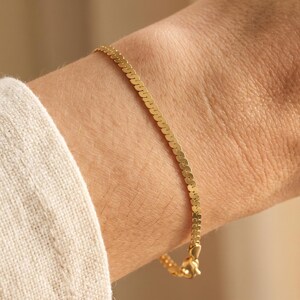 May include: A thin gold chain bracelet featuring a flat, interlocking herringbone link design. The bracelet has a polished, reflective surface and is secured with a small metal clasp visible at the end of the chain, resting against the skin.