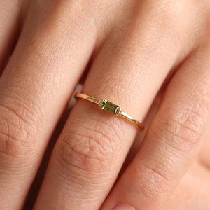 May include: A thin gold band ring featuring a single, small, rectangular, baguette-cut green gemstone set horizontally in the center. The metal band is smooth and polished, showcasing a minimalist design with a vibrant, translucent olive-green stone.