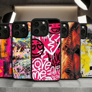 May include: A collection of black phone cases with vibrant graffiti art designs. The cases feature abstract patterns, hearts, and text like "Love Me," in a mix of pink, yellow, and white against black backgrounds. The cases are displayed on a gray surface.
