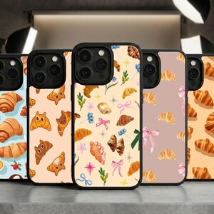 May include: Five phone cases with black frames, each featuring a unique design. The cases showcase illustrations of croissants, some with cat faces, and other pastries, set against various pastel backgrounds, including blue, peach, and beige.