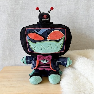 Pode incluir: Um brinquedo de pelúcia robô com uma cabeça preta e um rosto semelhante a uma tela com detalhes em vermelho, verde e azul. O brinquedo tem 25 cm de altura e 18 cm de largura, pesando 164 g. Possui uma gravata borboleta e está em uma pequena mesa de madeira.