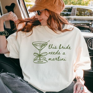 May include: Off-white crew neck t-shirt with a green martini glass graphic and the text "this bride needs a martini". The shirt is made of a soft material and is suitable for casual wear.