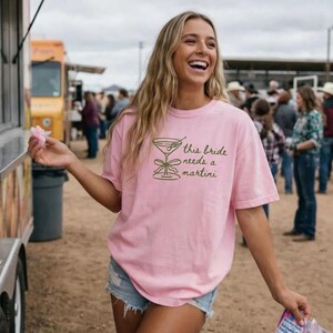 May include: A light pink t-shirt with the text "this bride needs a martini" in green. The shirt features a green martini glass graphic. The person is wearing denim shorts and is smiling.