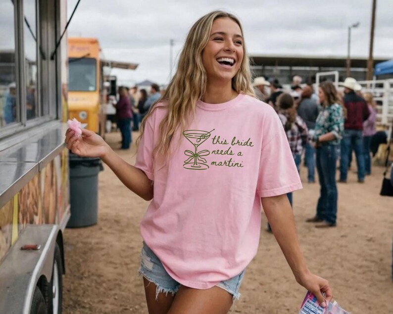 May include: A light pink t-shirt with the text "this bride needs a martini" in green. The shirt features a green martini glass graphic. The person is wearing denim shorts and is smiling.