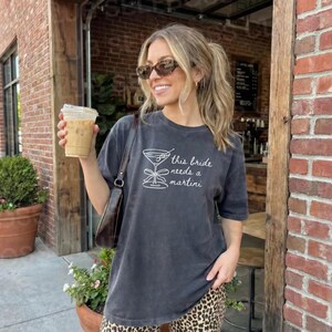 May include: A dark gray t-shirt with the text "this bride needs a martini" in white. The shirt features a martini glass graphic. The person in the image is wearing sunglasses and holding a beverage.