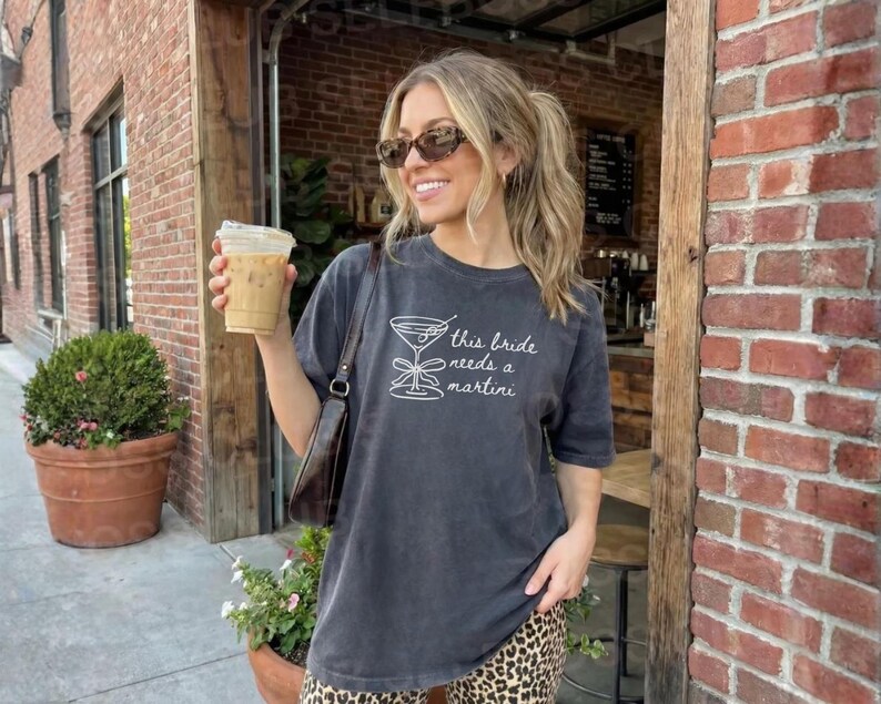 May include: A dark gray t-shirt with the text "this bride needs a martini" in white. The shirt features a martini glass graphic. The person in the image is wearing sunglasses and holding a beverage.