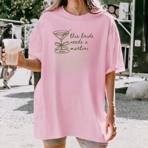 May include: A light pink t-shirt with the text "this bride needs a martini" and a martini glass graphic in green. The shirt is a relaxed fit, suitable for casual wear.