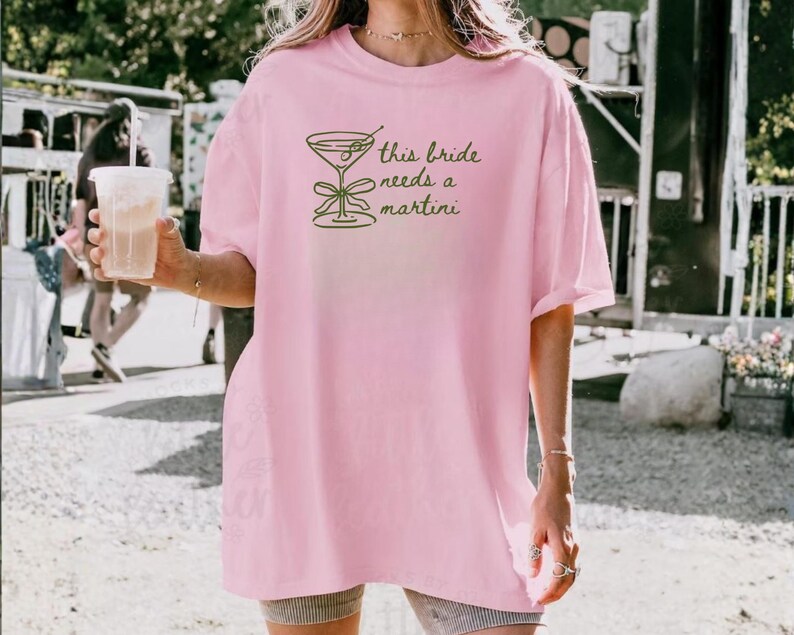 May include: A light pink t-shirt with the text "this bride needs a martini" and a martini glass graphic in green. The shirt is a relaxed fit, suitable for casual wear.