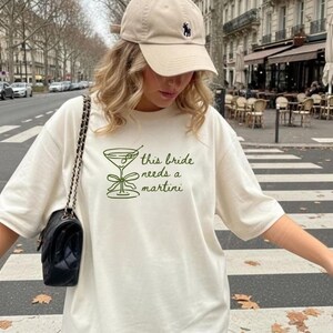 May include: Cream-colored t-shirt with the text "this bride needs a martini" in green. The shirt features a martini glass graphic. A black purse with a chain strap and a beige baseball cap are also visible.