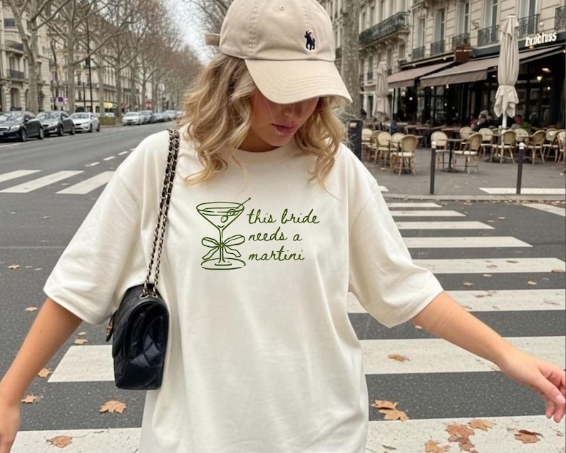 May include: Cream-colored t-shirt with the text "this bride needs a martini" in green. The shirt features a martini glass graphic. A black purse with a chain strap and a beige baseball cap are also visible.