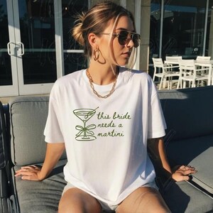 May include: White t-shirt with the text "this bride needs a martini" in green, along with a martini glass graphic. The shirt is worn by a person with sunglasses and gold jewelry.